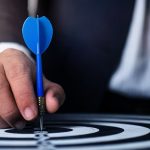 Close-up of a man's hand holding a blue dart on a bullseye, symbolizing precision and focus.